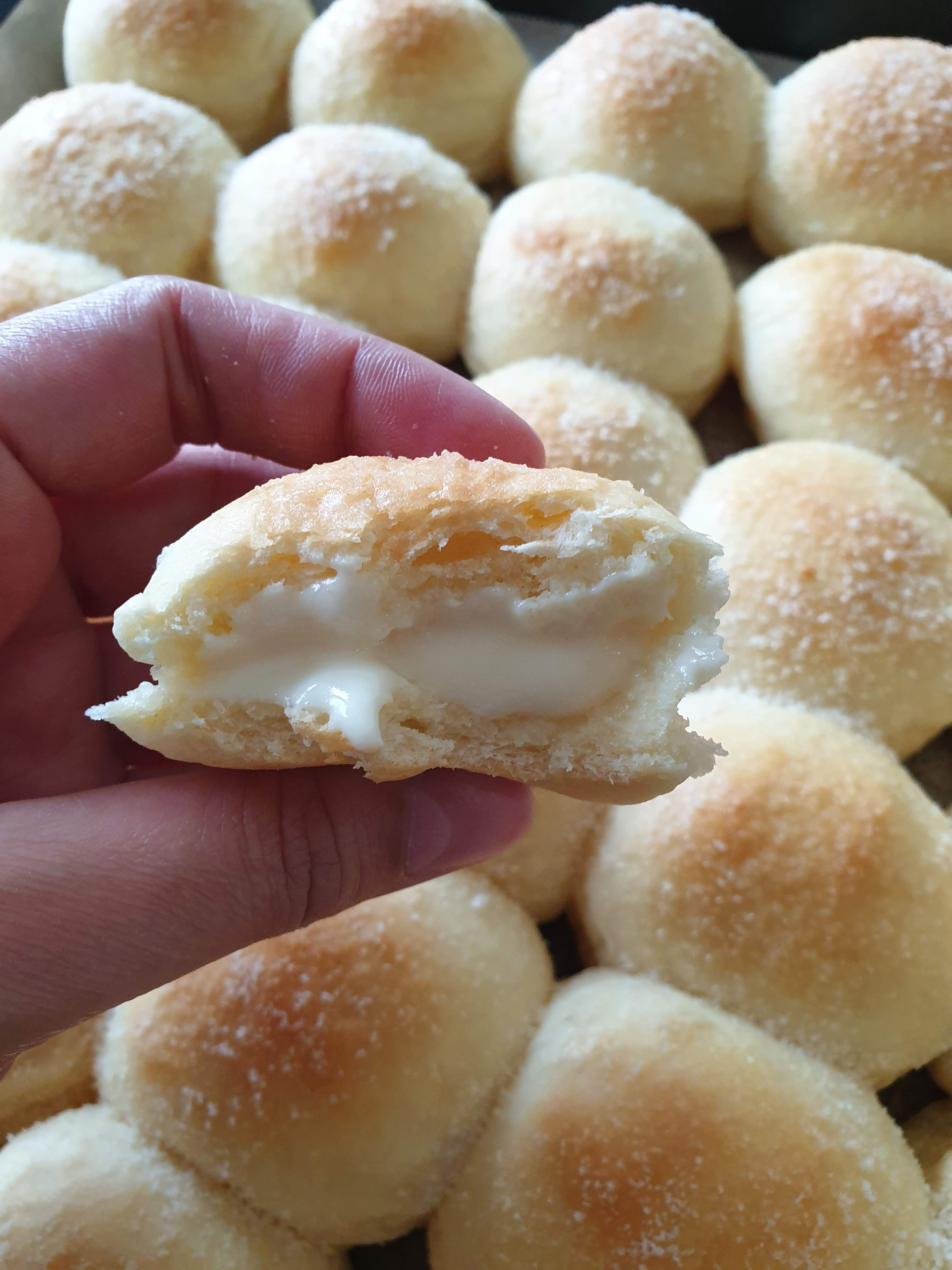 Mão segurando um pãozinho mordido, no fundo, vários pãezinhos.